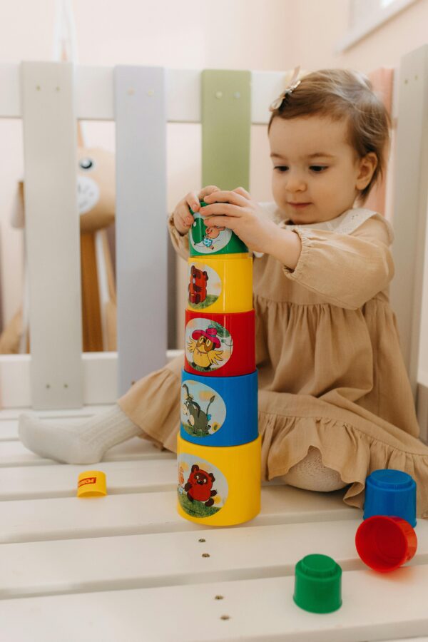 A toddler girl sits on a white picket floor, happily playing with colorful stacking cups in a light-filled room.