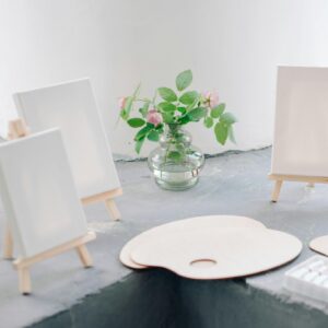 Art supplies setup with blank canvases on easels, paint palettes, and a jar of flowers.