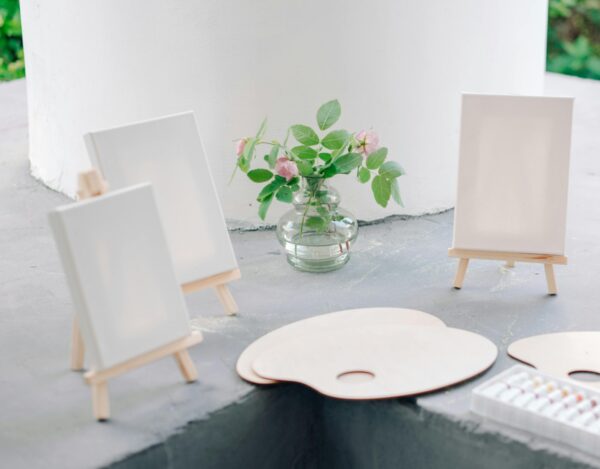 Art supplies setup with blank canvases on easels, paint palettes, and a jar of flowers.